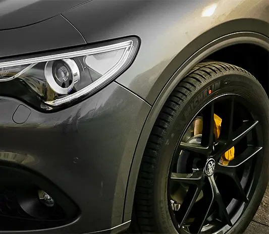 Close-up of an Alfa Romeo SUV's headlight and black alloy wheel with yellow brake caliper, serviced at Exotic Auto Services workshop.